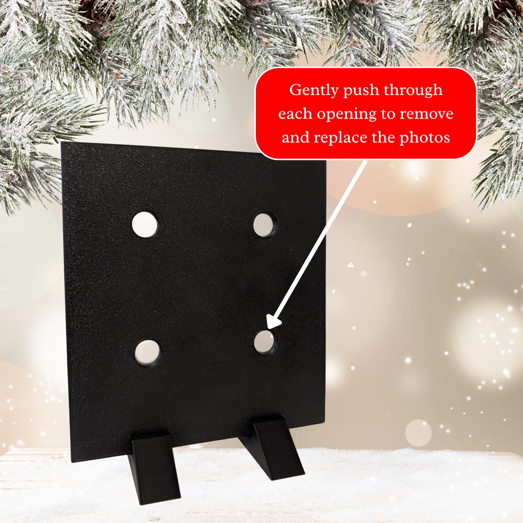 A black square photo frame display with four circular openings, shown on a snowy surface with pine branches overhead and a red instructional text bubble explaining how to remove and replace the photos.