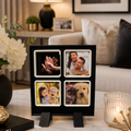 A black four-photo magnet frame with white borders and small black easel feet stands on a marble surface in a living room. The four slots contain sample photos: hands, a couple, children, and a dog.