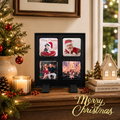 A black four-photo Christmas picture frame displayed on a wooden shelf in a decorated holiday setting, with a lit candle, pinecones, a small Christmas tree, books, and a house-shaped ornament nearby. The photos inside show festive holiday scenes and people.