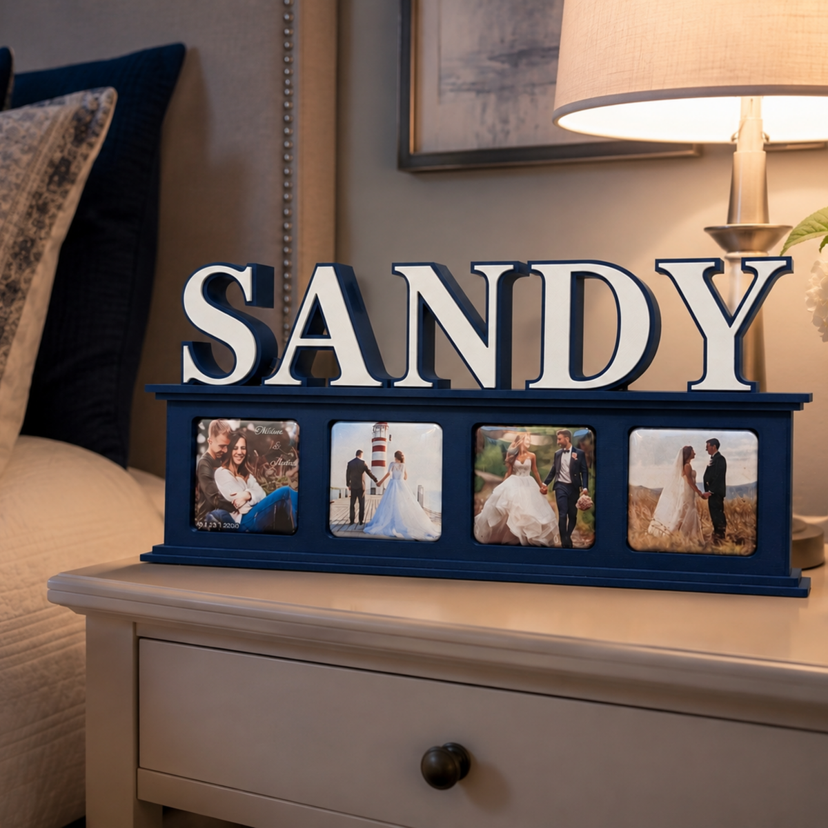A personalized navy blue photo frame display with the name 'SANDY' in large white letters, displayed on a bedside table next to a lit lamp and bed.