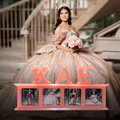 A woman in an elaborate champagne ball gown with a tiara standing in front of a staircase, holding a bouquet of pink flowers. In the foreground is a coral-pink customized photo frame spelling 'KAE' with four photo magnets showing her in different outfits.