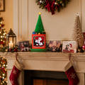 A festive Christmas mantel display featuring a small green artificial tree in a red and green box with a photo frame on top, surrounded by stockings, holiday lights, cards, and a small white tree against a warm beige wall.