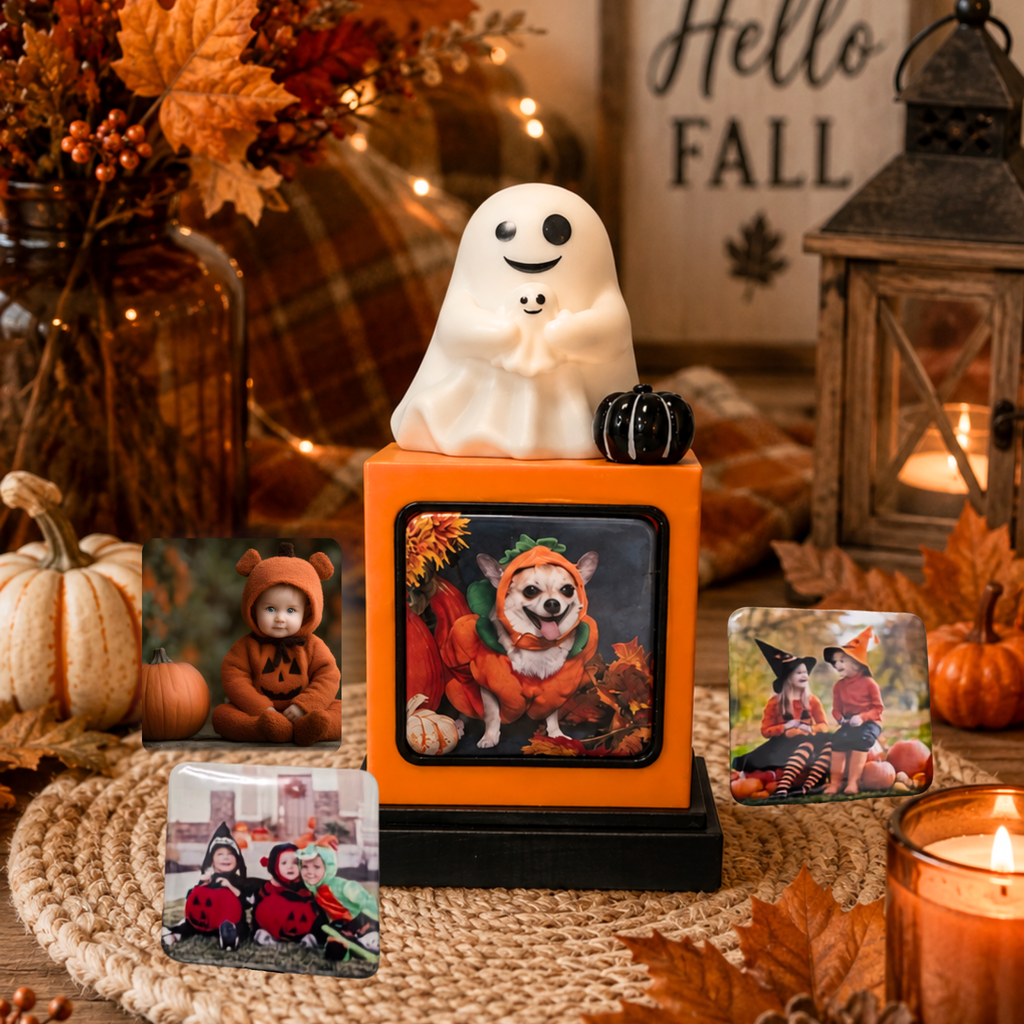 A Halloween-themed photo display featuring a Mommy and Baby ghost figurine on top of an orange cube with a black base, surrounded by fall decorations, small pumpkins, candles, and several printed custom photos arranged on a woven mat.