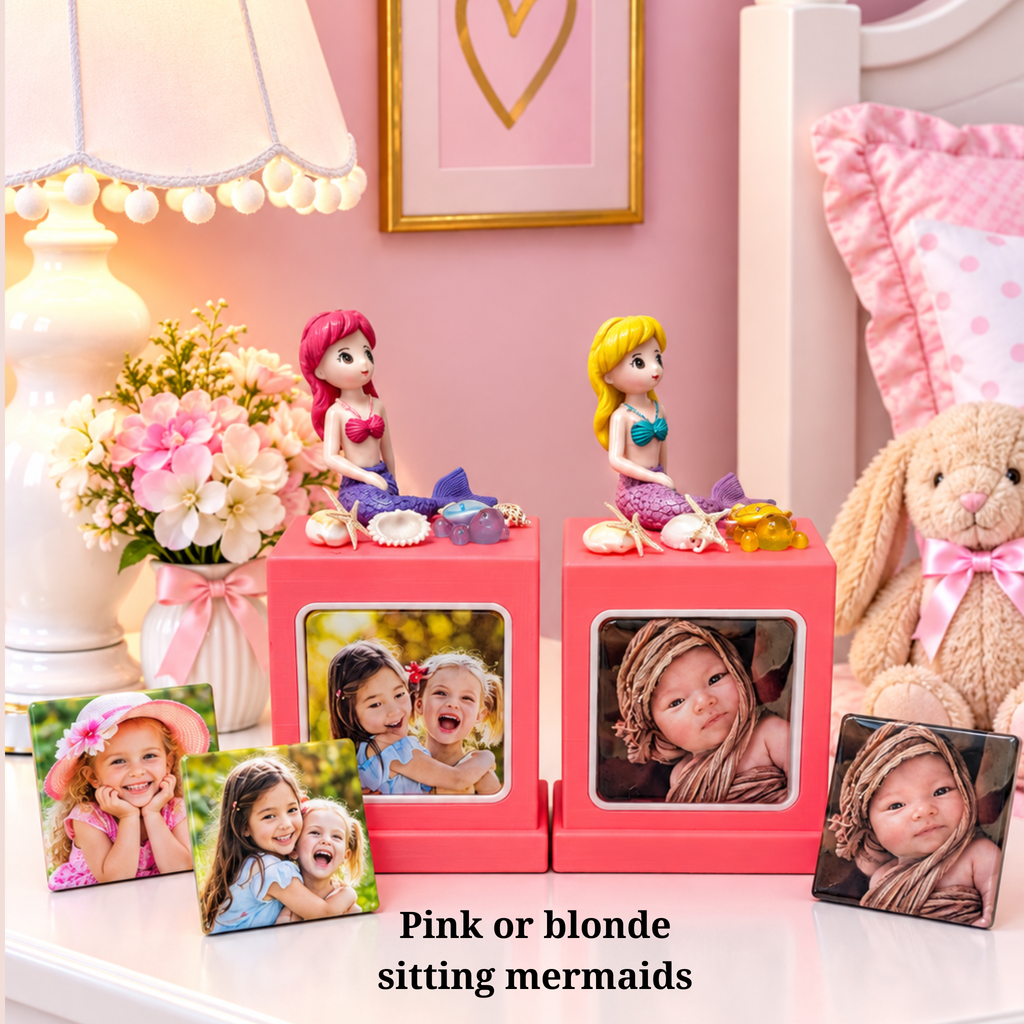 A pink memory cube photo frame stand set styled in a nursery, featuring two square pink frames with mermaid figurines on top and two custom photo inserts. The text overlay reads 'Pink or blonde sitting mermaids'.