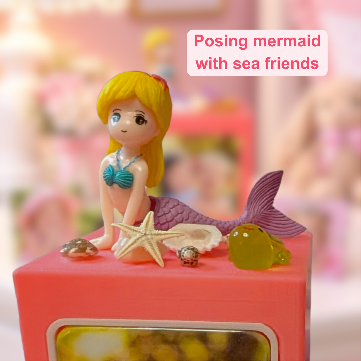 A mermaid figurine with blonde hair, a teal top, and a purple tail sits on a pink memory cube with sea friends. The background is softly blurred pink, and a text box reads 'Posing mermaid with sea friends'.
