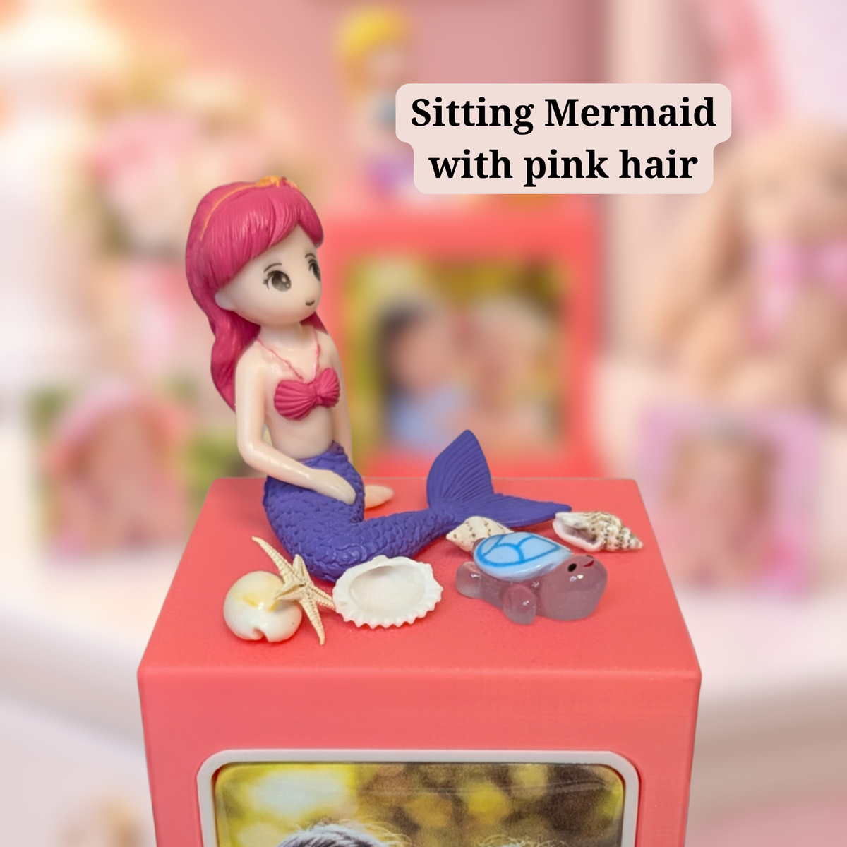 A mermaid figurine with pink hair sits on a pinking memory cube photo display, surrounded by small seashells, a starfish, and a turtle. The background is softly blurred, and a text label reads 'Sitting Mermaid with pink hair'.