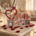 A decorative tabletop photo frame display with a large red heart outline and the word LOVE in red letters on top of a white rectangular photo magnet frame holding four photos magnets. The piece is styled on a distressed cream dresser with lace, scattered red rose petals, and a softly blurred bedroom background.